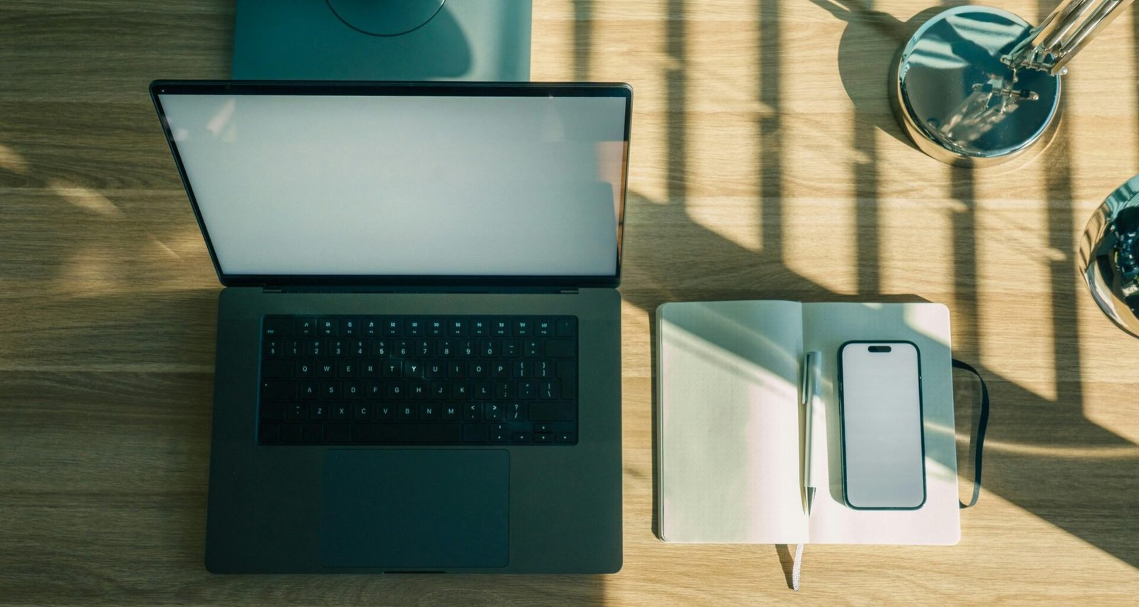 Laptop, Handy und Notizbuch liegen nebeneinander auf einem hölzernen Schreibtisch, bereit zum Arbeiten.