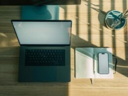 Laptop, Handy und Notizbuch liegen nebeneinander auf einem hölzernen Schreibtisch, bereit zum Arbeiten.