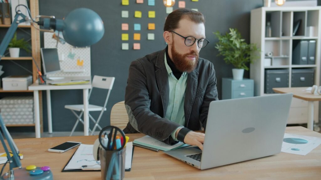 Ein Mann sitzt an einem Schreibtisch in einem modernen Büro und arbeitet an seinem Laptop, umgeben von technischer Ausstattung.