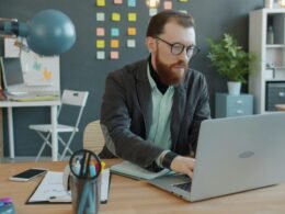 Ein Mann sitzt an einem Schreibtisch in einem modernen Büro und arbeitet an seinem Laptop, umgeben von technischer Ausstattung.