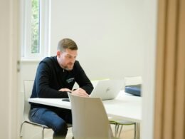 Ein Mann sitzt an einem weißen Tisch und arbeitet an einem Laptop. Im Hintergrund unscharfe Arbeitsmaterialien.