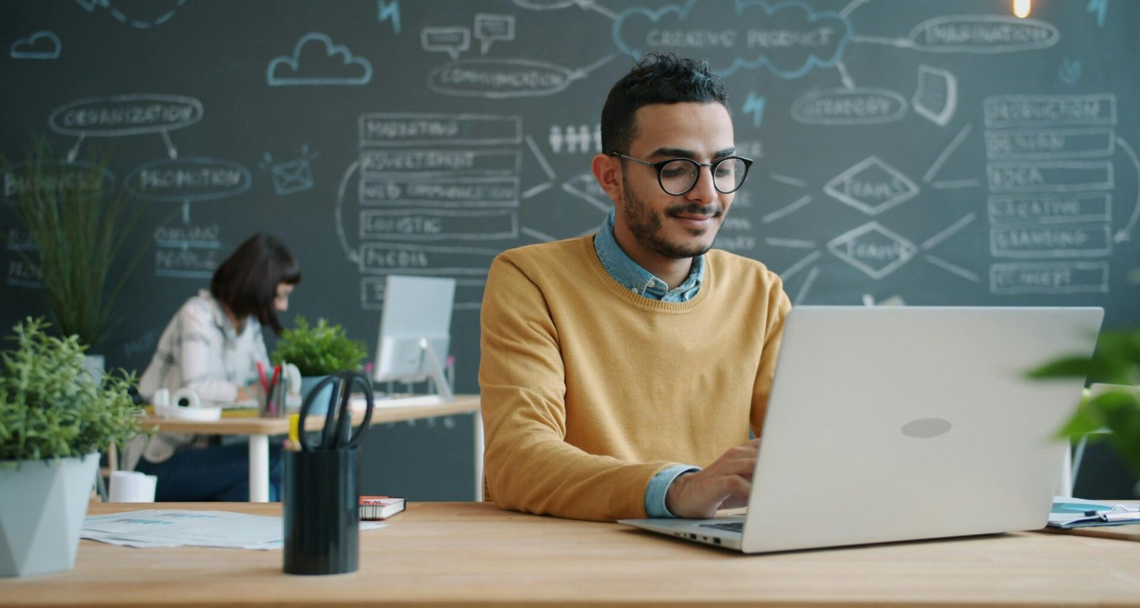 Mann sitzt an einem Schreibtisch und arbeitet mit einem Laptop in einem modernen Büro mit Whiteboard im Hintergrund.