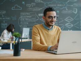 Mann sitzt an einem Schreibtisch und arbeitet mit einem Laptop in einem modernen Büro mit Whiteboard im Hintergrund.