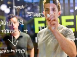 Mann schreibt mit Marker auf eine transparente Glaswand und plant Strategien oder Konzepte in einem modernen Büro