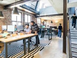 Stehender Mann bespricht sich mit sitzendem Kollegen am Schreibtisch, während dieser am Computer arbeitet