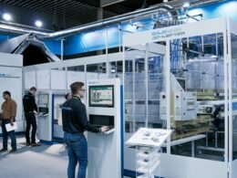 Ein Mann arbeitet am Computer, daneben tauscht sich eine Gruppe Männer in einem Büro aus