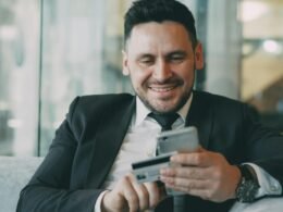 Ein Geschäftsmann im Anzug hält lächelnd ein Smartphone in der einen und eine Kreditkarte in der anderen Hand.