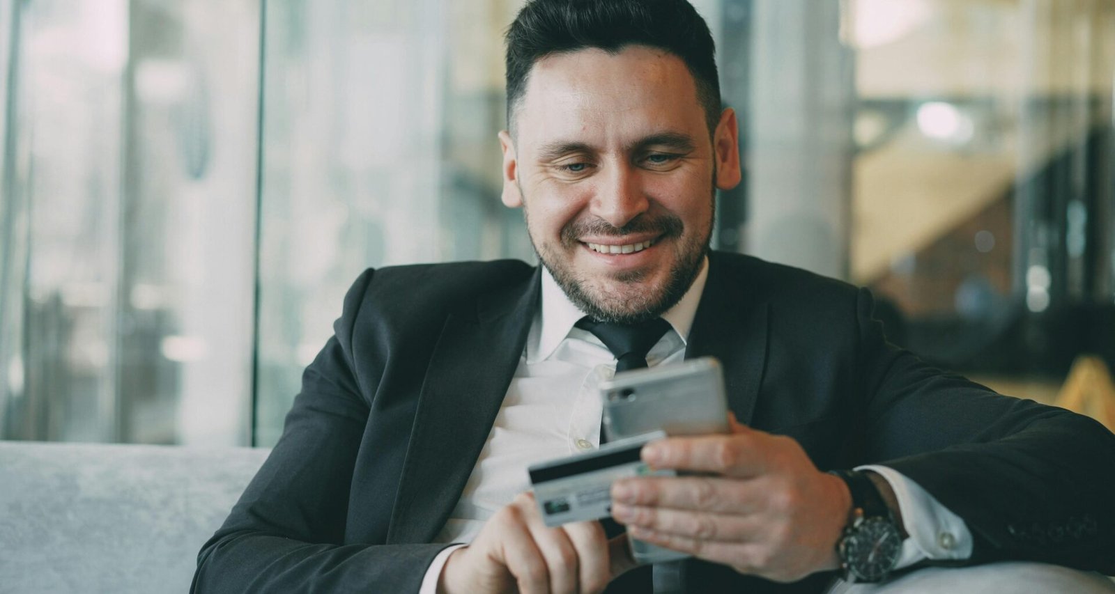 Mann im Anzug lächelt und hält ein Smartphone sowie eine Kreditkarte in der Hand, moderner Geschäftskontext