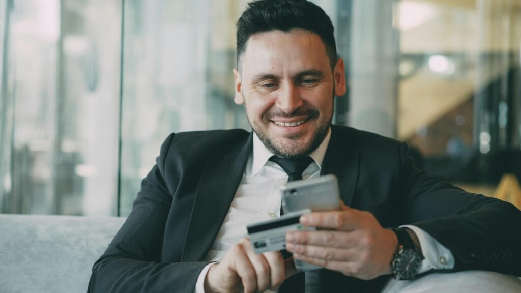 Mann im Anzug hält Smartphone und Kreditkarte in der Hand, zeigt dabei ein Lächeln, Fokus auf digitale Nutzung