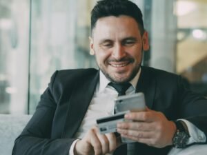 Mann im Anzug lächelt, während er ein Smartphone und eine Kreditkarte hält – Symbolbild für Online-Zahlungen