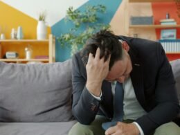 Mann im Anzug sitzt auf einem Sofa und stützt den Kopf in die Hände, fotografiert im Innenraum