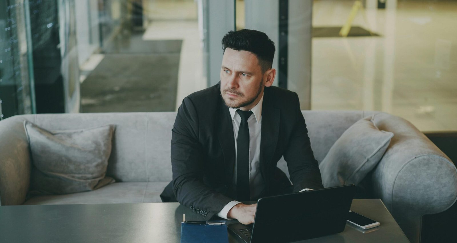 Mann im Anzug sitzt auf einem Sofa und arbeitet mit einem Laptop, moderne Büroumgebung im Hintergrund.