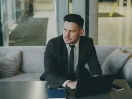 Mann im Anzug sitzt auf einem Sofa und arbeitet mit einem Laptop, moderne Büroumgebung im Hintergrund.