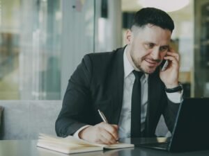 Mann im Anzug telefoniert und schreibt in ein Notizbuch an einem Schreibtisch im Büro