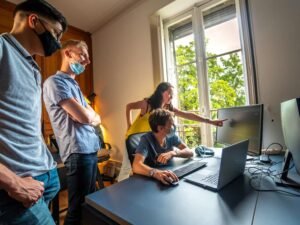 Zwei Männer stehen nebeneinander, einer im blauen Hemd, einer im gelben Polohemd, und unterhalten sich in einem Innenraum.