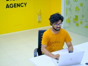 Mann in orangefarbenem Crewneck-T-Shirt sitzt auf schwarzem Stuhl vor neutralem Hintergrund.