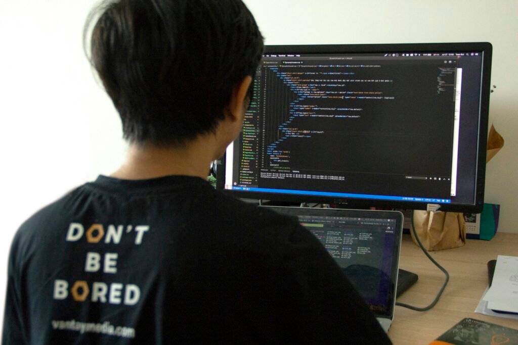 Mann in schwarzem Shirt arbeitet an einem Laptop und nutzt zusätzlich einen großen Flachbildschirm am Schreibtisch