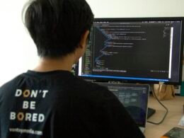 Mann in schwarzem Shirt sitzt an Schreibtisch und arbeitet mit Laptop und Flachbildschirm-Monitor