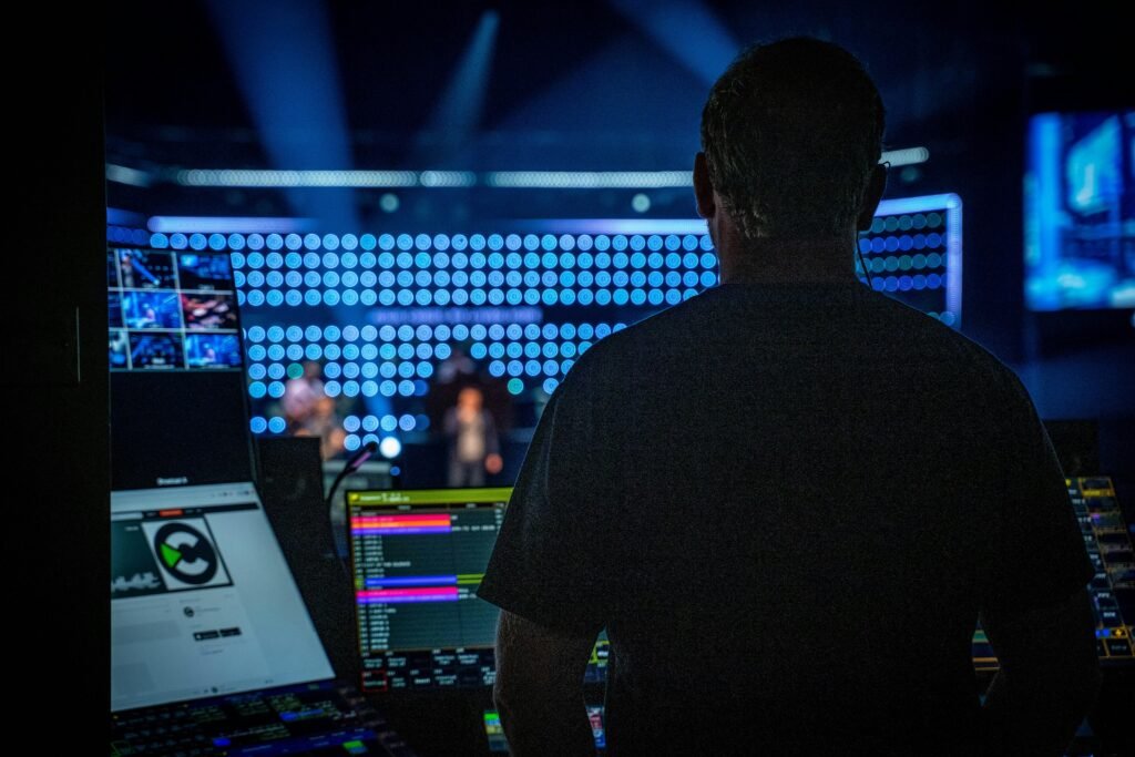 Ein Mann in schwarzem Shirt steht vor einem professionellen Audiomischpult und überwacht die Tonregler während einer Produktion.