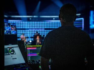 Ein Mann in schwarzem Shirt steht vor einem professionellen Audiomischpult und überwacht die Tonregler während einer Produktion.