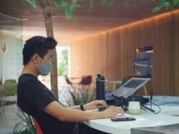 Ein Mann im schwarzen T-Shirt sitzt an einem Schreibtisch vor einem aufgeklappten Laptop und arbeitet konzentriert.