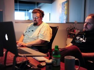 Mann in weißem T-Shirt sitzt auf braunem Stuhl vor neutralem Hintergrund, alltägliche Büroszene