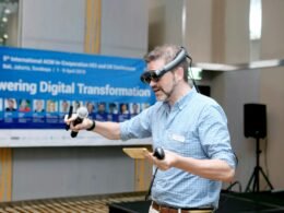 Mann in blau-weiß kariertem Hemd hält ein schwarz-braunes Mikrofon in der Hand, aufgenommen in neutraler Umgebung.