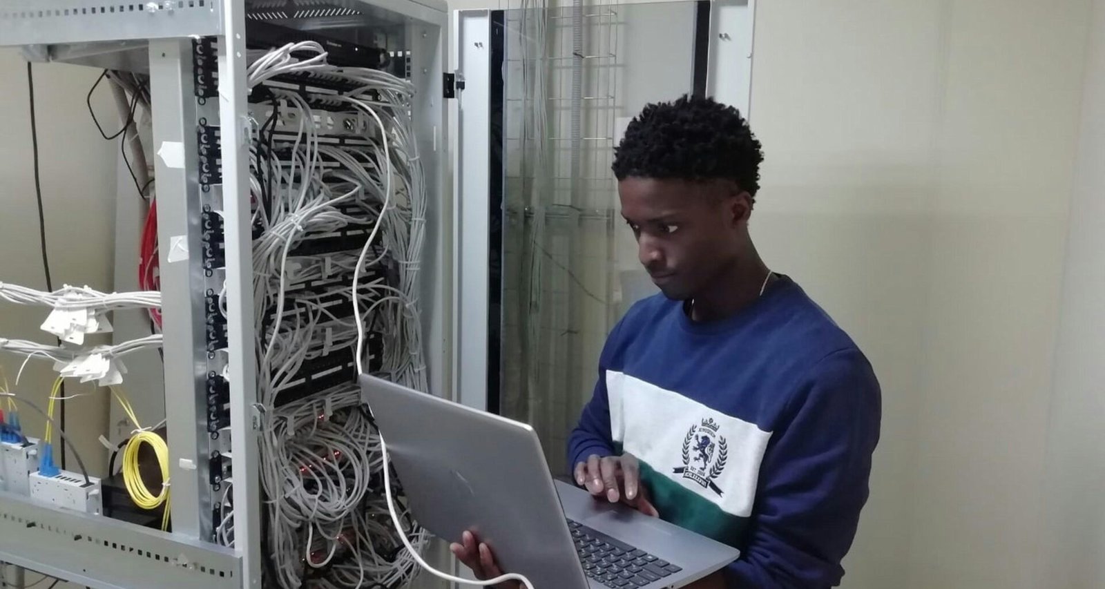 Ein Mann mit blauem Pullover arbeitet fokussiert an einem silbernen MacBook-Laptop an einem Schreibtisch.