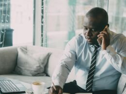Mann im Büroumfeld mit Hemd und Krawatte, telefonierend am Schreibtisch sitzend, moderner Arbeitsplatz mit Unterlagen