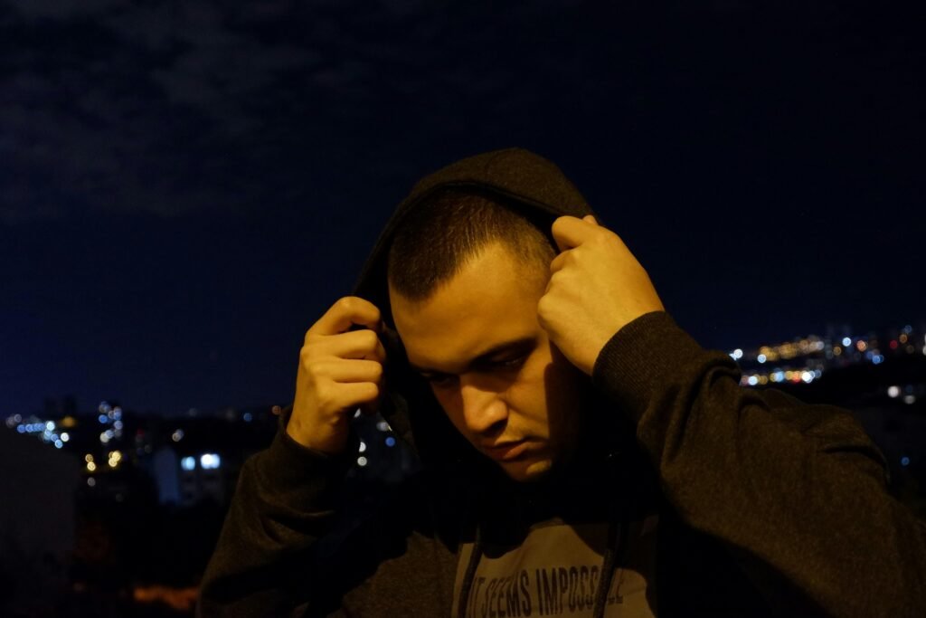 Ein Mann trägt einen schwarzen Hoodie und verdeckt teilweise sein Gesicht mit den Händen, fotografiert vor neutralem Hintergrund.