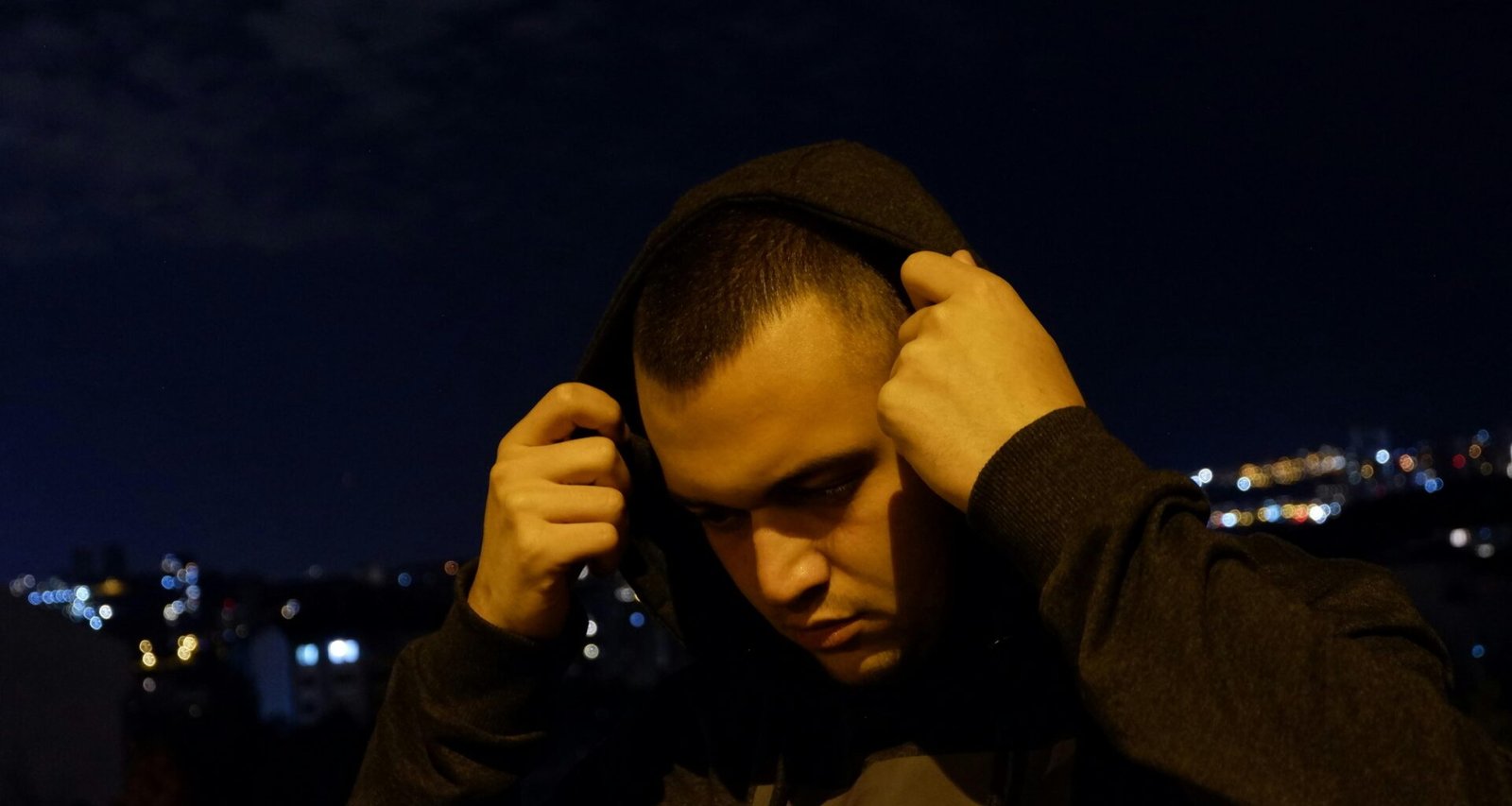 Ein Mann trägt einen schwarzen Hoodie und verdeckt teilweise sein Gesicht mit den Händen, fotografiert vor neutralem Hintergrund.