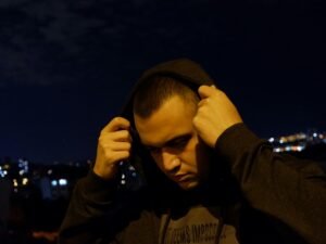 Ein Mann trägt einen schwarzen Hoodie und verdeckt teilweise sein Gesicht mit den Händen, fotografiert vor neutralem Hintergrund.