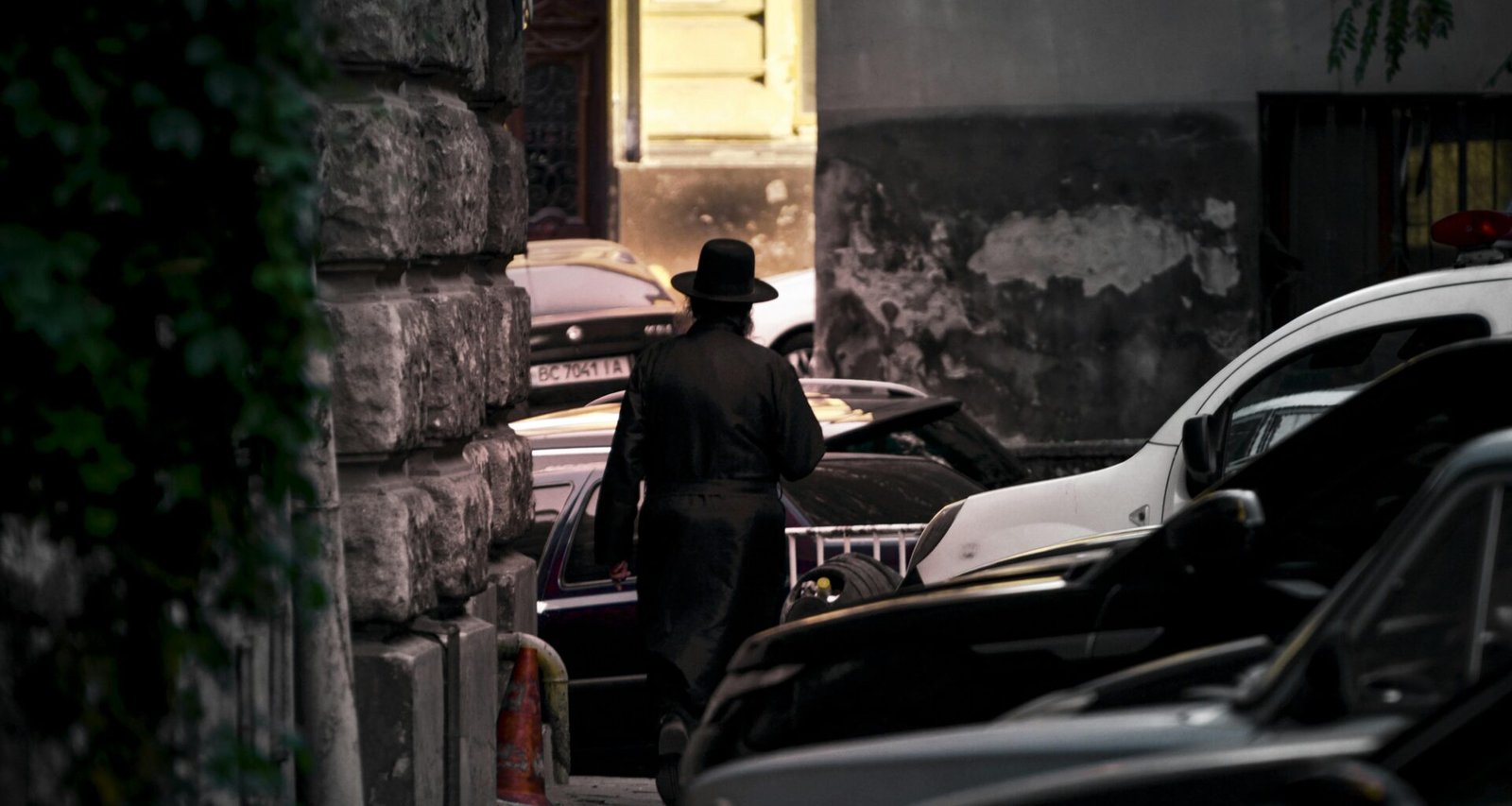 Ein Mann in schwarzer Jacke und braunem Hut steht tagsüber neben einem schwarzen Auto auf einer Straße