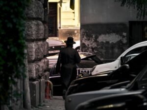 Ein Mann in schwarzer Jacke und braunem Hut steht tagsüber neben einem schwarzen Auto auf einer Straße