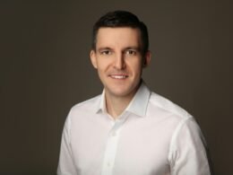 Lächelnder Mann mit weißem Hemd vor einem einfarbigen grauen Hintergrund, neutraler Business-Look