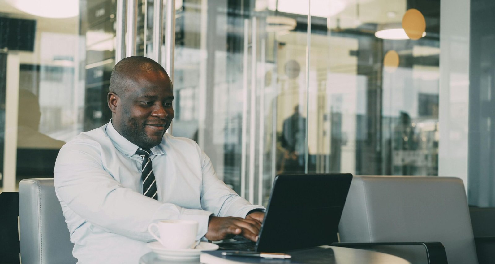 Ein Mann sitzt an einem Tisch in einem Café und arbeitet lachend an seinem Laptop, modernes Ambiente im Hintergrund.