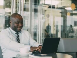 Ein Mann sitzt an einem Tisch in einem Café und arbeitet lachend an seinem Laptop, modernes Ambiente im Hintergrund.