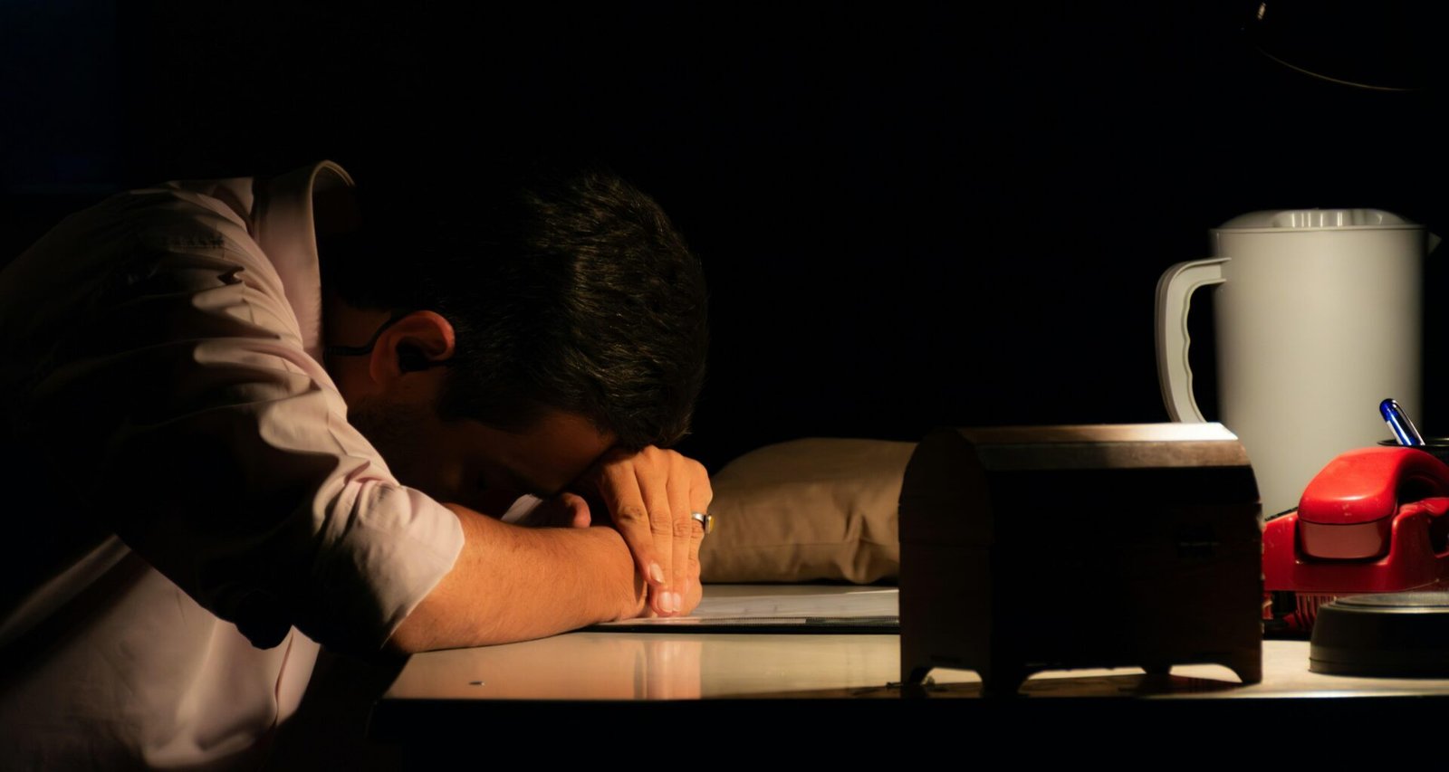 Ein Mann sitzt in einem dunklen Raum und legt den Kopf auf den Schreibtisch, umgeben von Technik und Computerbildschirmen.