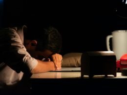 Ein Mann sitzt in einem dunklen Raum und legt den Kopf auf den Schreibtisch, umgeben von Technik und Computerbildschirmen.