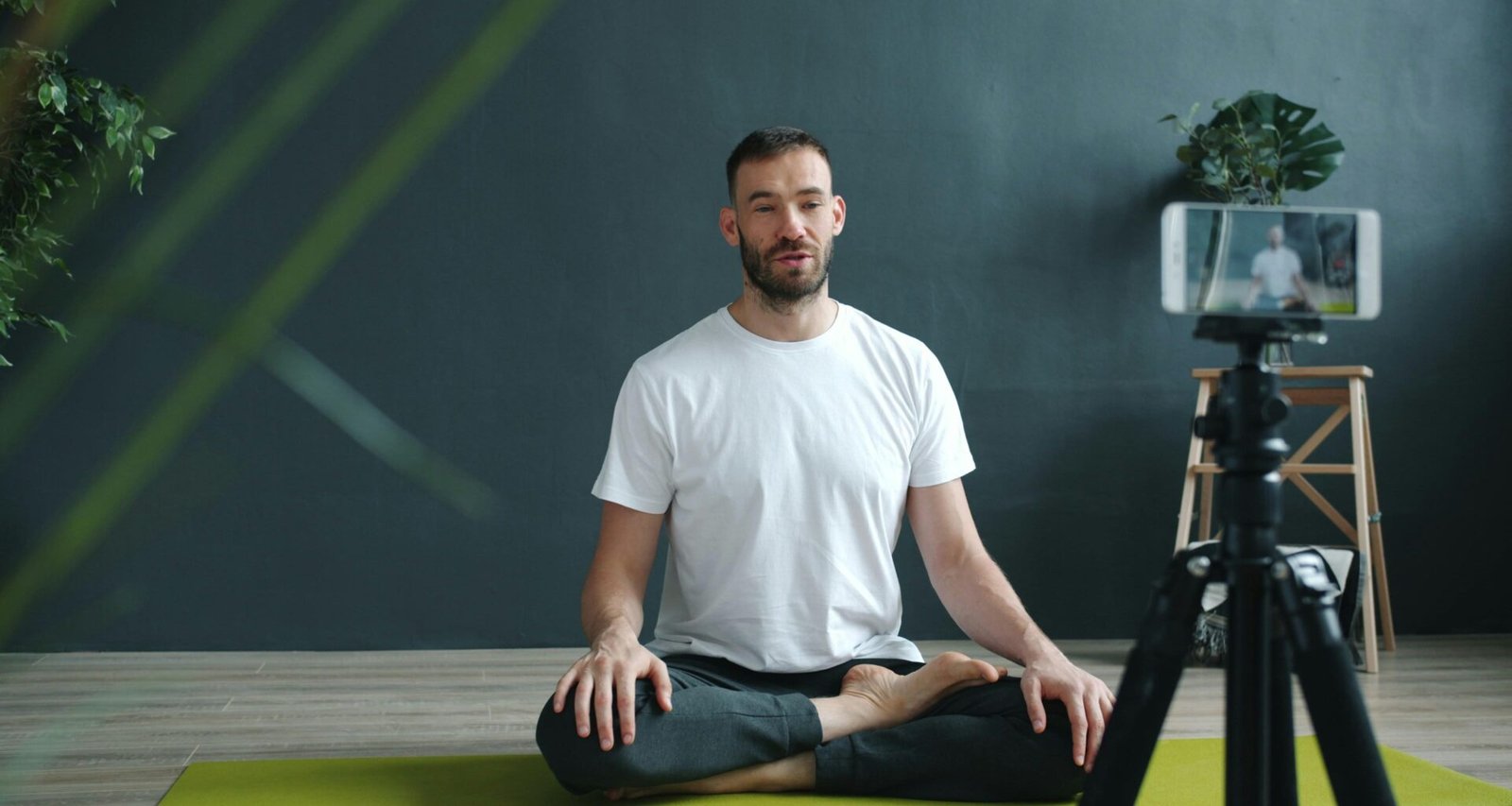 Mann sitzt meditierend auf einer Yogamatte und filmt sich mit seinem Smartphone für eine digitale Meditationssession.