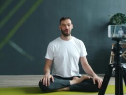 Mann sitzt meditierend auf einer Yogamatte und filmt sich mit seinem Smartphone für eine digitale Meditationssession.