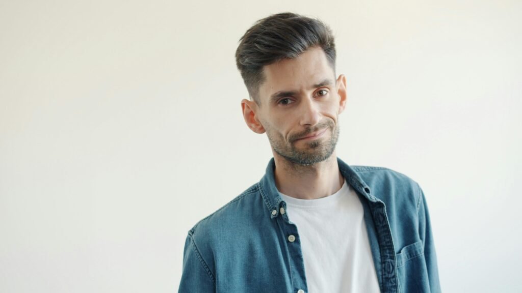 Mann mit Bart in blauem Jeanshemd und weißem T-Shirt vor neutralem Hintergrund, frontal im Bild