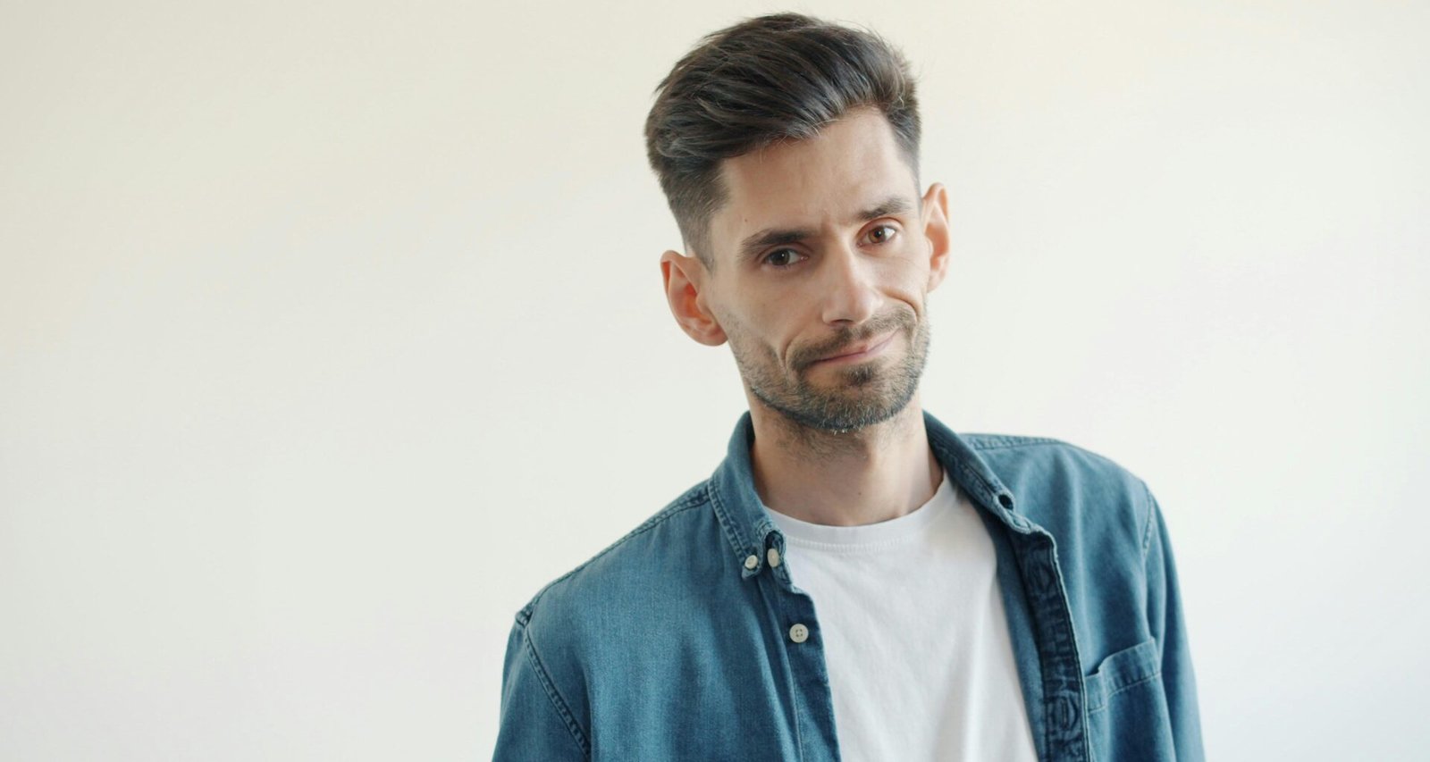 Mann mit Bart in blauem Jeanshemd und weißem T-Shirt vor neutralem Hintergrund, frontal im Bild