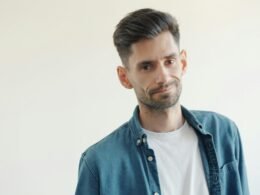 Mann mit Bart in blauem Jeanshemd und weißem T-Shirt vor neutralem Hintergrund, frontal im Bild