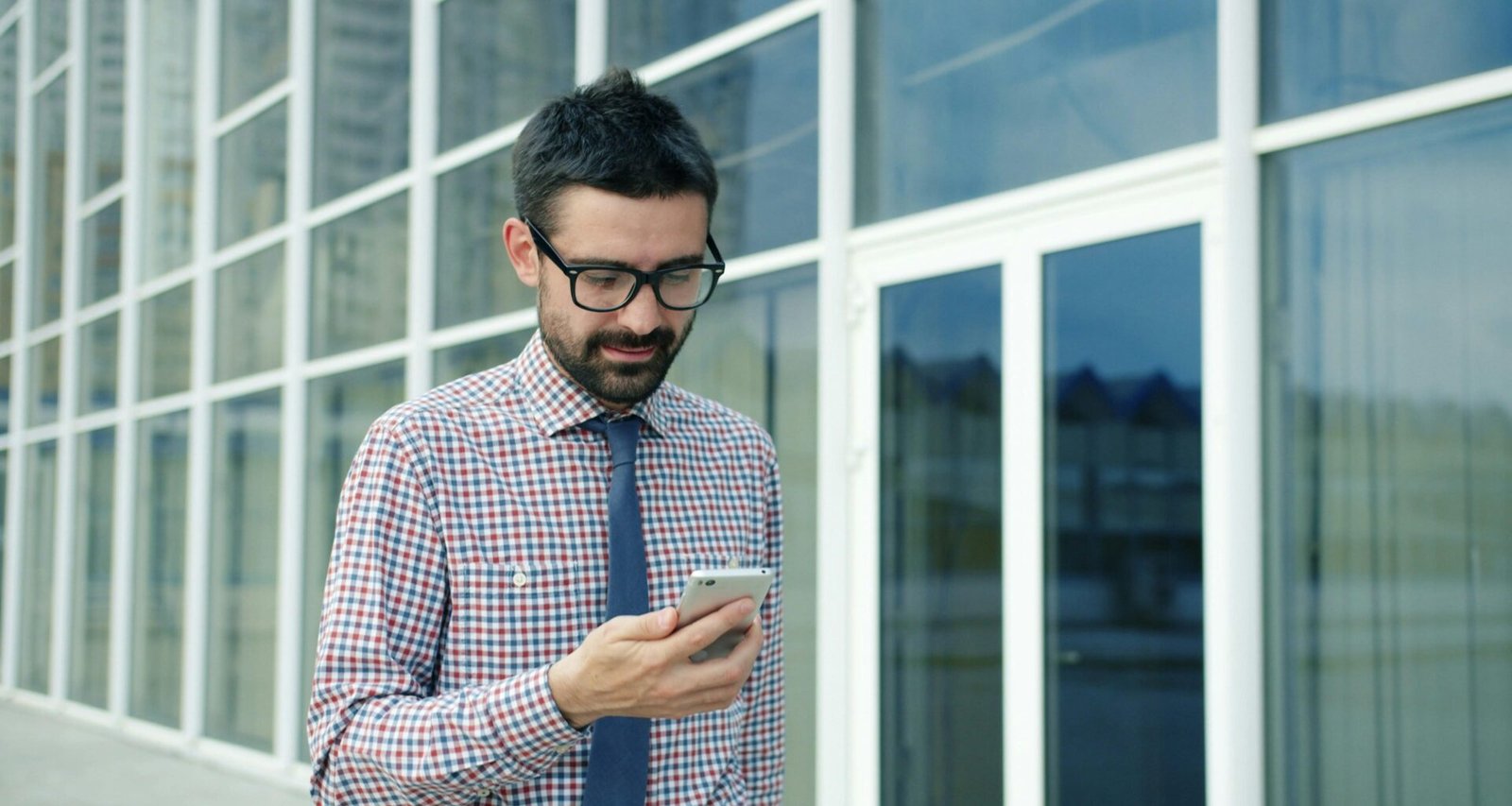 Mann mit Brille steht draußen vor einem modernen Gebäude und schaut konzentriert auf sein Smartphone