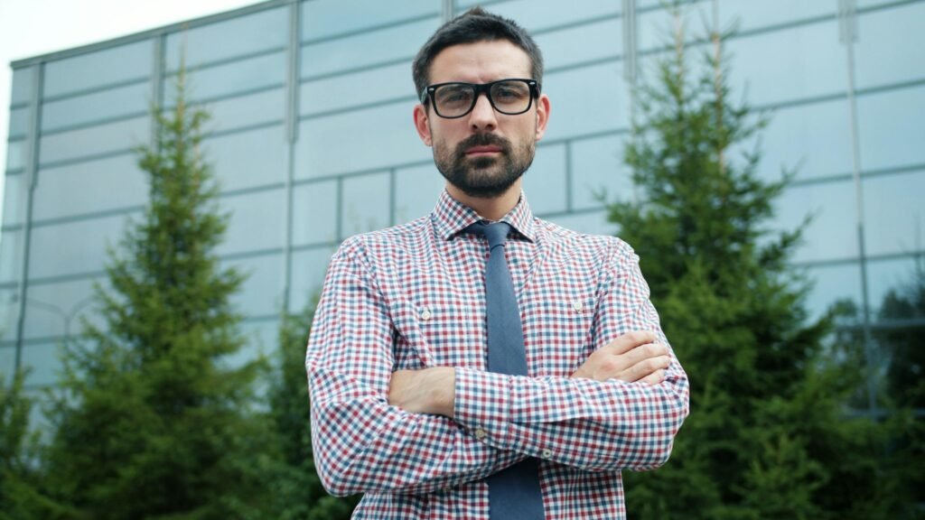 Mann mit Brille und verschränkten Armen steht draußen vor einem modernen Gebäude, neutraler Hintergrund