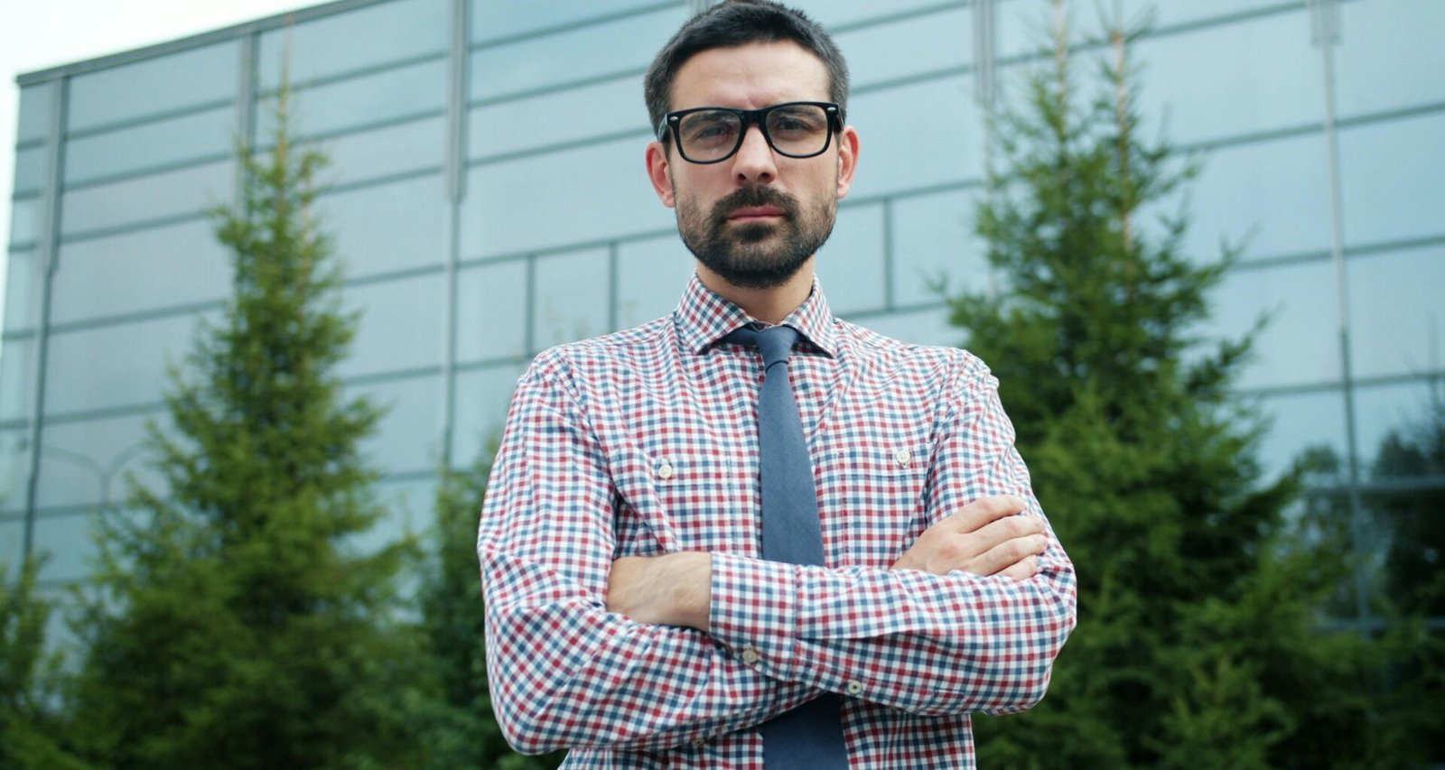 Mann mit Brille und verschränkten Armen steht draußen vor einem modernen Gebäude, neutraler Hintergrund