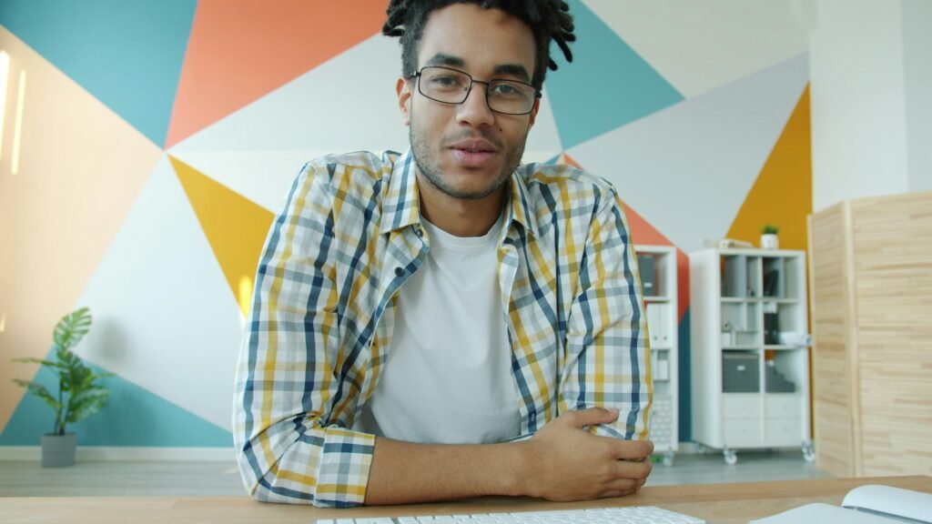 Mann mit Dreadlocks und Kopfhörern bei einem Videocall am Schreibtisch vor Laptop und Notizbuch