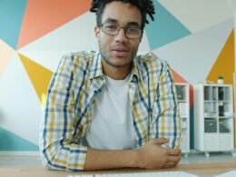 Mann mit Dreadlocks und Kopfhörern bei einem Videocall am Schreibtisch vor Laptop und Notizbuch