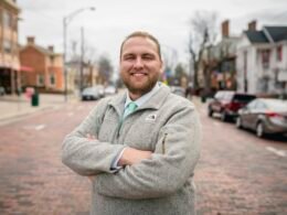 Mann mit kurzgeschnittenem Bart, trägt eine graue Jacke und steht vor einem neutralen Hintergrund. Geeignet für Business-Themen.